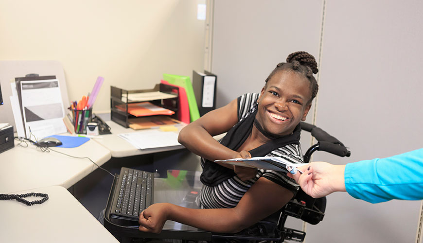 A person in an electric wheelchair eraches back from her desk, smiling as they take a file from a colleague