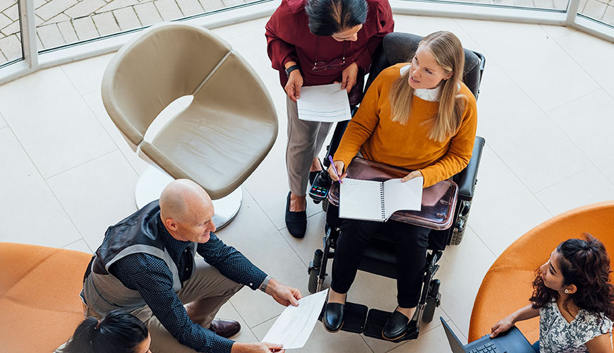 people sitting in a circle with laptops and notebooks, one of whom is in a wheelchair