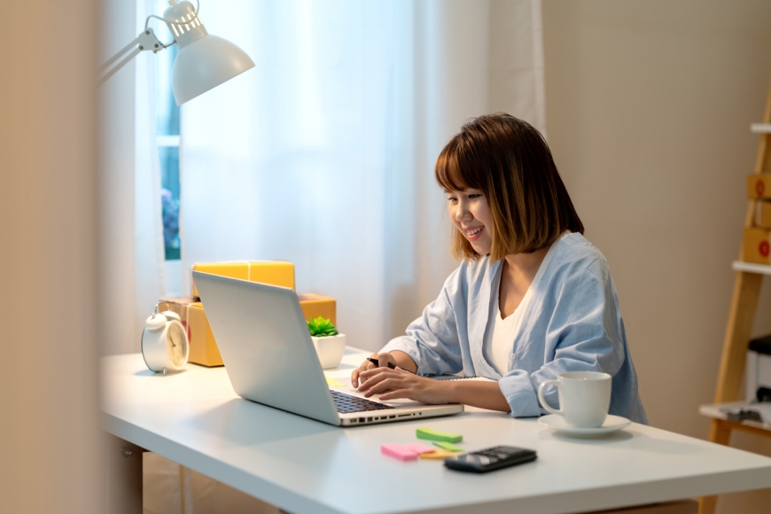 A young woman dressed casually sits at a desk in a home office, looking at a laptop computer. 