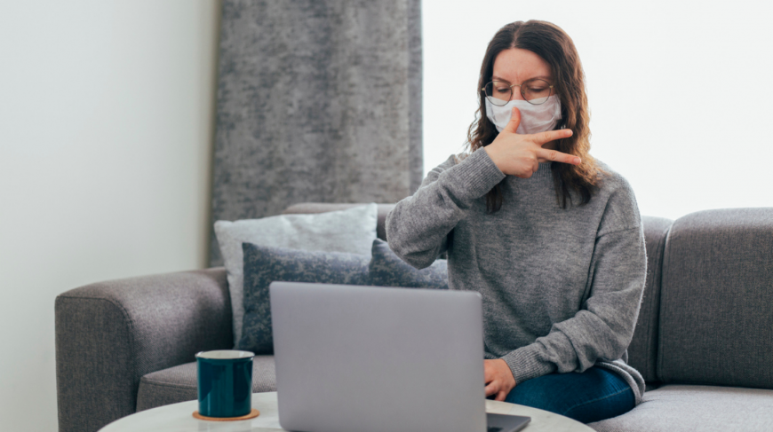 A young woman sits on a sofa looking at a laptop, using sign language and wearing a mask.