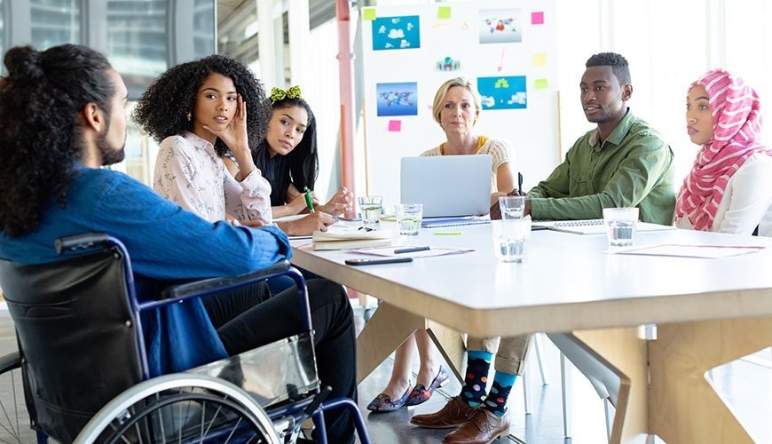 A group of colleagues sitting around a table from various ethnicities and ages.  One man is sitting in a wheelchair.