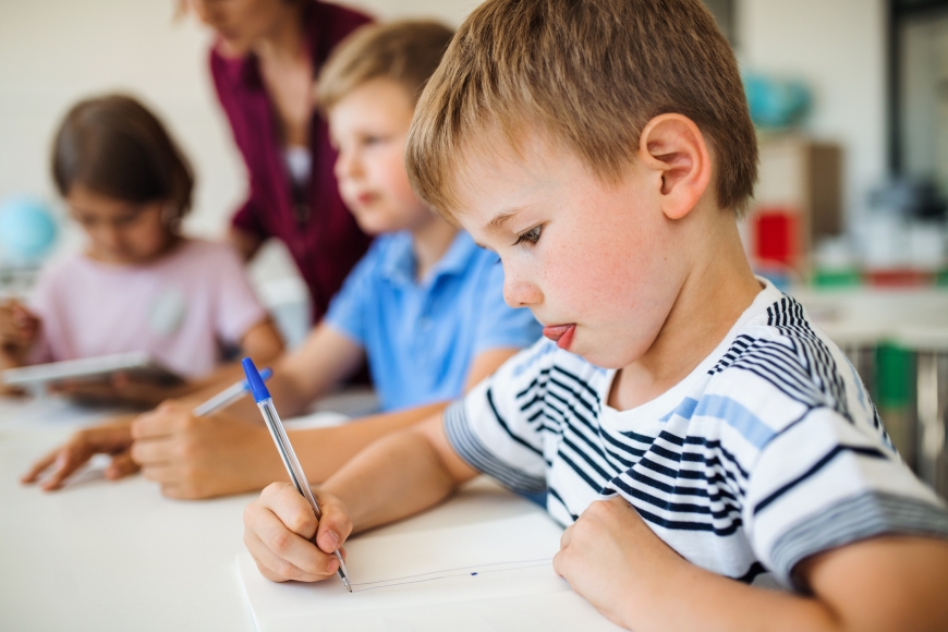 Three children in a classroom, a teacher visible in the background.