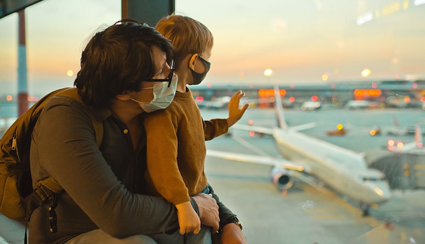 Father and small child at airport, looking out the window at waiting planes. Both are wearing covid masks. 