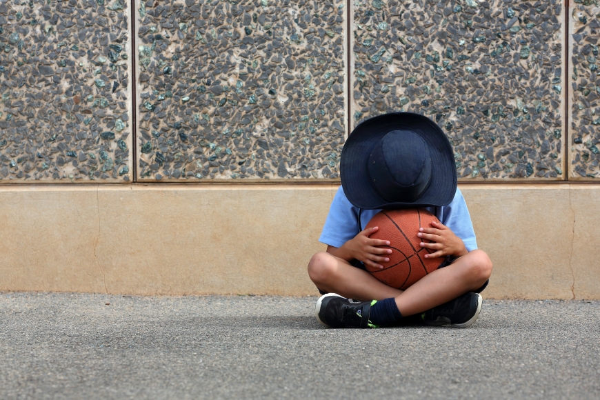 School boy sitting cross legged hiding face in a basketball.