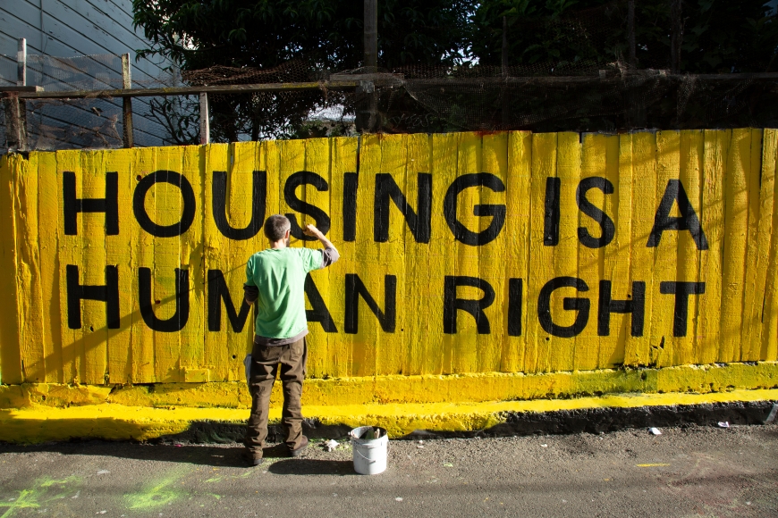 man paints 'housing is a human right' on yellow wooden fence