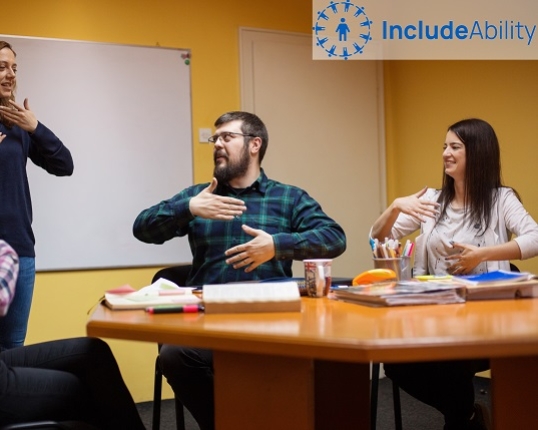 People in a workplace meeting communicate with sign-language, the Includeability logo is overlaid in the top-right corner of the image.