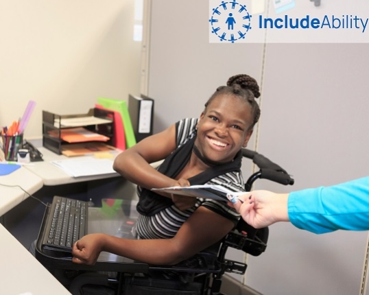 A person in at a wheelchair smiles from their desk and hands a document to a colleague, the Includeability logo is overlaid in the top-right corner of the image.