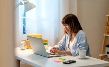 A young woman dressed casually sits at a desk in a home office, looking at a laptop computer. 