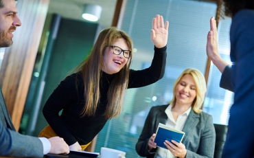 A person with Down Syndrome holds her hand up high-fiving colleagues in an office setting