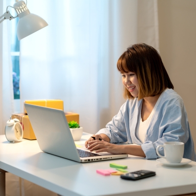 A young woman dressed casually sits at a desk in a home office, looking at a laptop computer. 