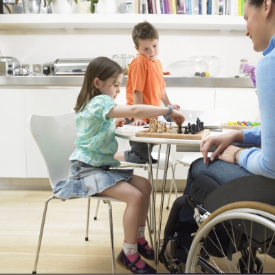 A woman using a wheelchair is at a table with two young children at home playing a game.