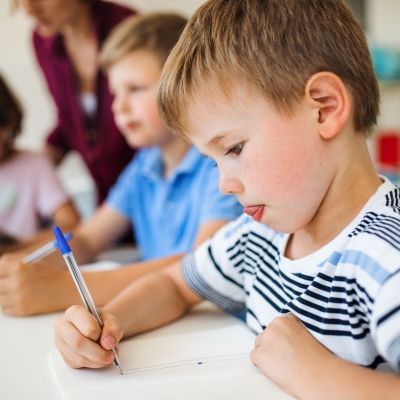 Three children in a classroom, a teacher visible in the background.