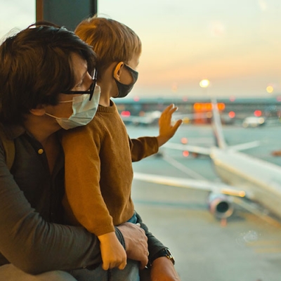 Father and small child at airport, looking out the window at waiting planes. Both are wearing covid masks. 