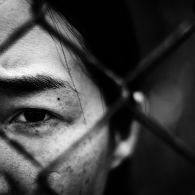 A sad woman looking this a chain-link fence.